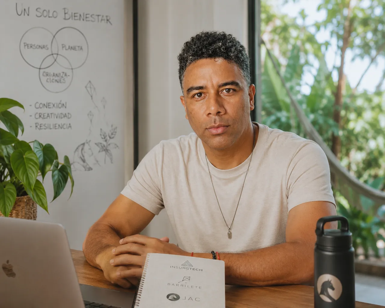 Eduardo Rengifo sentado a una mesa de madera; detrás, una pizarra con tres círculos que se cruzan —personas, planeta, organizaciones— bajo el título «Un solo bienestar», y sobre la mesa una libreta con los nombres Insurgtech, Barrilete y JAC